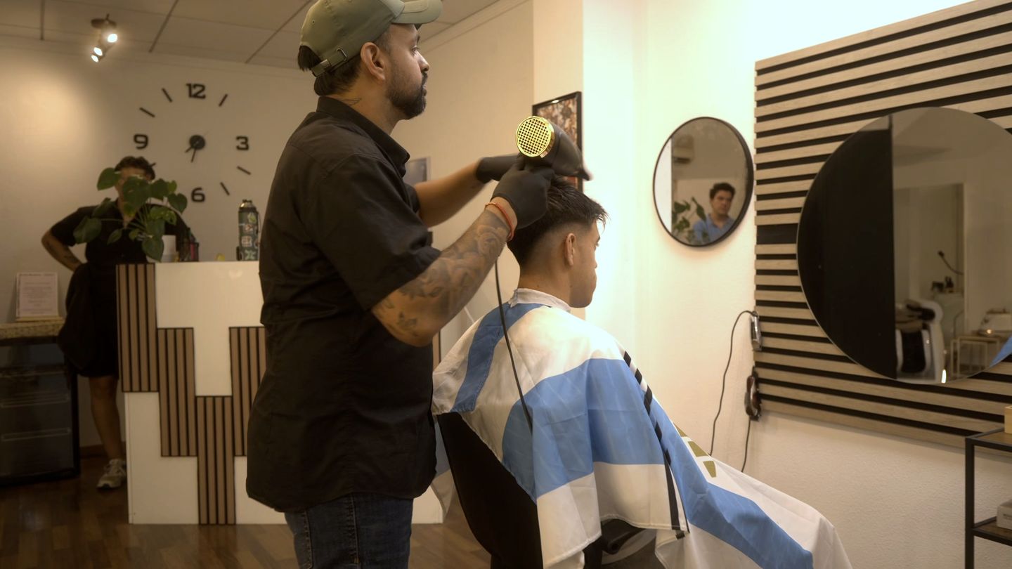 El peluquero seca el pelo a un cliente en el sillón, con el reloj, la recepción de listones y los espejos redondos del salón